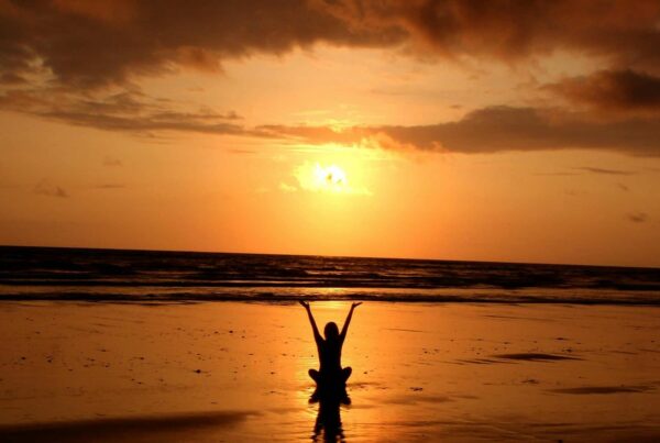 Woman at a beach at sunset sitting in the shallows with arms raised; she is balanced from her wellness treatment